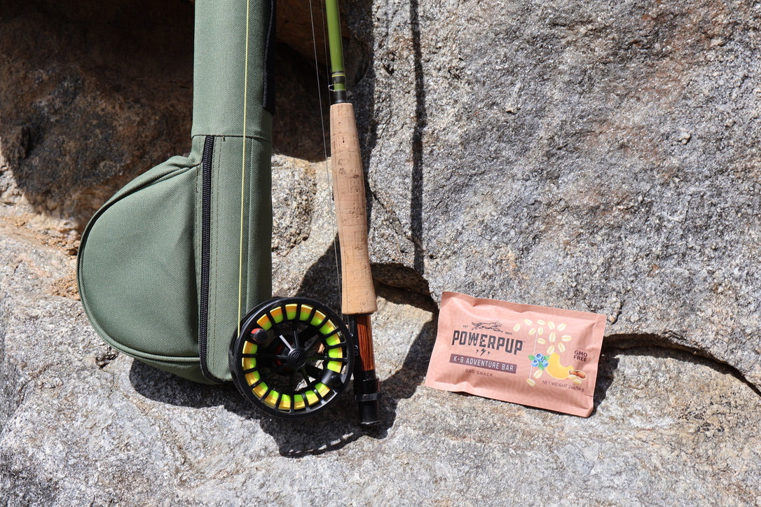 A fly fishing rod with a cork handle and black reel lies on a gray rock beside an olive green soft case and a pink POWERUP energy snack bar